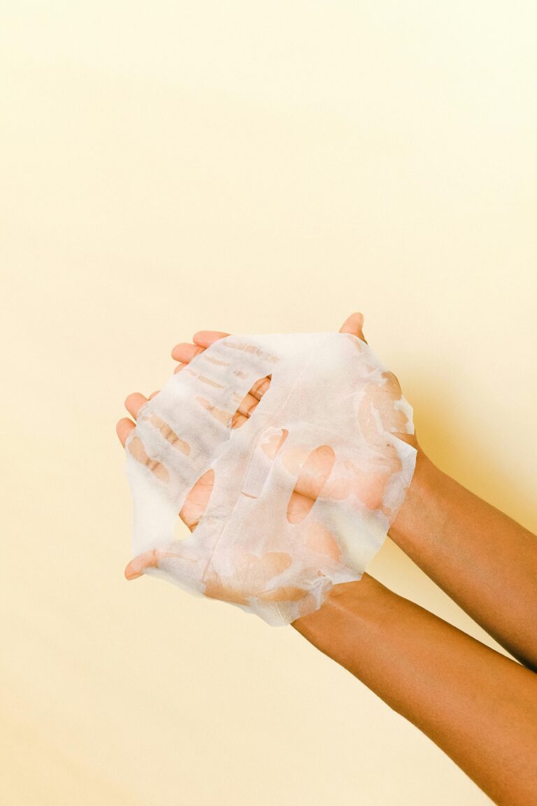 Close-up of hands holding a white sheet mask for skincare on a light background.